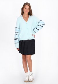 Ljusblå mjuk cardigan med V-ringning, fem knappar och mönstrade ärmar med hjärtan. Matchad med en plisserad svart kjol och vita sneakers.