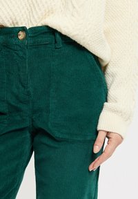 Pantalons en velours côtelé verts avec des nervures verticales, dotés d'une taille et d'une fermeture à bouton unique, associés à un pull en maille de couleur crème.