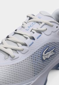 Λευκό και γαλάζιο αθλητικό παπούτσι με διχτυωτό επάνω μέρος, λευκά κορδόνια, το κείμενο "LACOSTE" στη γλώσσα και το λογότυπο κροκόδειλου στη πλευρά.