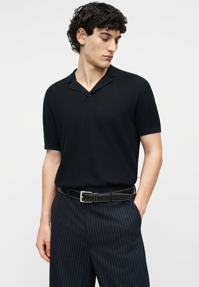 Άνδρας με σγουρά μαλλιά που φοράει μαύρο κοντομάνικο πλεκτό πόλο, μαύρη ζώνη και μπλε παντελόνι με ρίγες, ποζάροντας με το ένα χέρι στην τσέπη.