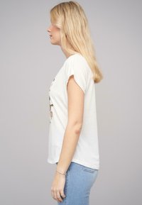 T-shirt blanc à manches courtes avec un col rond, ornée d'un motif floral dans des tons naturels. Le tissu semble doux et décontracté.