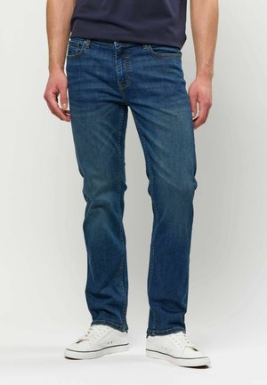 Mężczyzna w niebieskich prostych dżinsach i białych trampkach, stojący na jednolitym jasnym tle z rękami swobodnie opuszczonymi wzdłuż ciała.