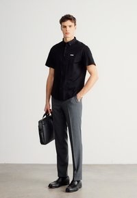 Μαύρο πουκάμισο με κοντό μανίκι και κουμπιά, με λογότυπο, συνδυασμένο με γκρι ραμμένα παντελόνια και μαύρα επίσημα παπούτσια, κρατώντας μια μαύρη δερμάτινη τσάντα χειρός.