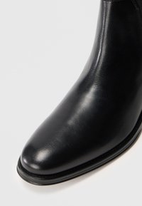 Bota de cuero negro liso con punta redonda y tacón bajo apilado, mostrada contra un fondo gris claro.