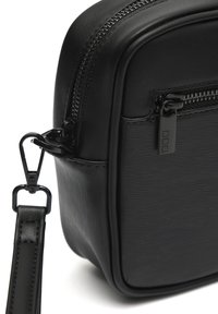 Bolso bandolera de cuero negro texturizado con cremallera visible, cierre de metal y clip de correa desmontable, mostrado en primer plano.