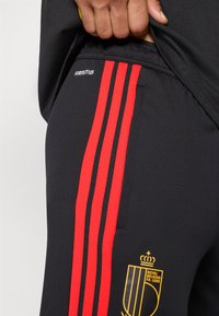 Calças de treino pretas com três riscas vermelhas de lado, com um logótipo em amarelo e o texto "FIRMOTION" perto da cintura.