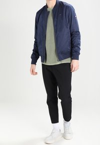 Granatowa kurtka bombowa z ściągaczami przy mankietach i kołnierzu, zielony sweter pod spodem, czarne tapered spodnie i białe sneakersy. Prosty, stonowany design.