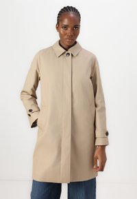 Imperméable beige à coupe droite, col boutonné, deux poches latérales et manches à revers. Tissu lisse avec un subtil éclat.