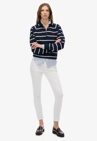 Un sweat à zip rayé bleu marine et blanc superposé sur une chemise col bleu clair, associé à un jean blanc et des mocassins foncés.
