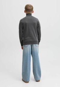 Un enfant aux cheveux blonds courts se tient de dos, portant un col roulé gris foncé et un jean bleu clair à jambes larges, avec des chaussures marron.