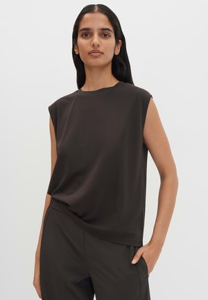 Femme aux longs cheveux noirs portant un haut noir sans manches et un pantalon noir, debout avec une main dans la poche devant un fond uni.