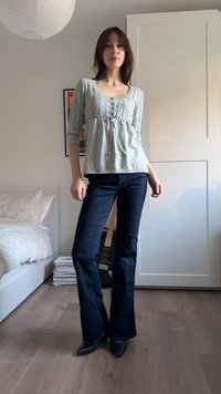Hellgrauer Pullover mit Zopfmuster und Rundhalsausschnitt, kombiniert mit dunkelblauen ausgestellten Jeans und schwarzen spitzen High Heels. Schlichter Schlafzimmerhintergrund.