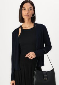 Cardigan marine en matériau léger, avec une fermeture à boutons et des poignets côtelés, associé à une jupe plissée noire et un sac à main.