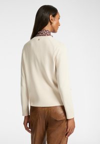 Cremfarbener, gerippter Pullover mit rundem Halsausschnitt, getragen über einem lila gemusterten Hemd. Auffällige Nähte und ein kleines Logodetail auf der Rückseite.