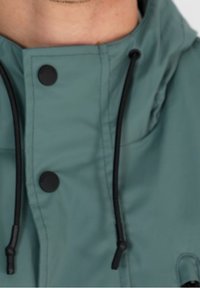 Zielona wodoodporna kurtka z czarnymi zatrzaskami i czarnymi sznurkami w okolicy szyi, noszona przez osobę.