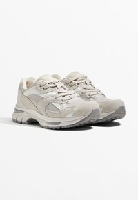 A világosszürke sportcipők háló és velúrbőr anyagok keverékével, lekerekített formájával, párnázott bokarészével és texturált talpával rendelkeznek a tapadás érdekében.