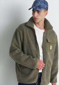 Giacca in pile verde oliva con texture morbida, zip frontale, colletto alto e tasca sul petto con foderato arancione. Abbinata a un berretto blu.