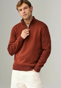 Svetr v rezavé barvě s vysokým límcem a polo zipem, s žebrovanými manžetami a lemem, nošený přes světlo zbarvenou košili, doplněný o krémové kalhoty.