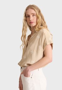Chemise beige à manches courtes en tissu léger, ornée de motifs floraux brodés blancs et d'une coupe décontractée. Associée à un pantalon blanc.
