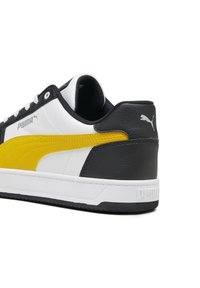 Bílé tenisky s černými a žlutými akcenty, s texturovaným koženým svrškem, gumovou podrážkou a brandingem Puma na boku a patě.