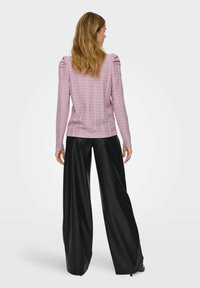 Kvinne med skulderlangt hår iført en rosa blonde langermet topp og vide svarte skinnbukser, stående vendt bort fra kameraet mot en hvit bakgrunn.