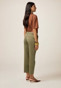 Camicia marrone a maniche corte, pantaloni a gamba larga verde oliva e scarpe slingback burgundy con punta affilata. Texture lisce e linee pulite.