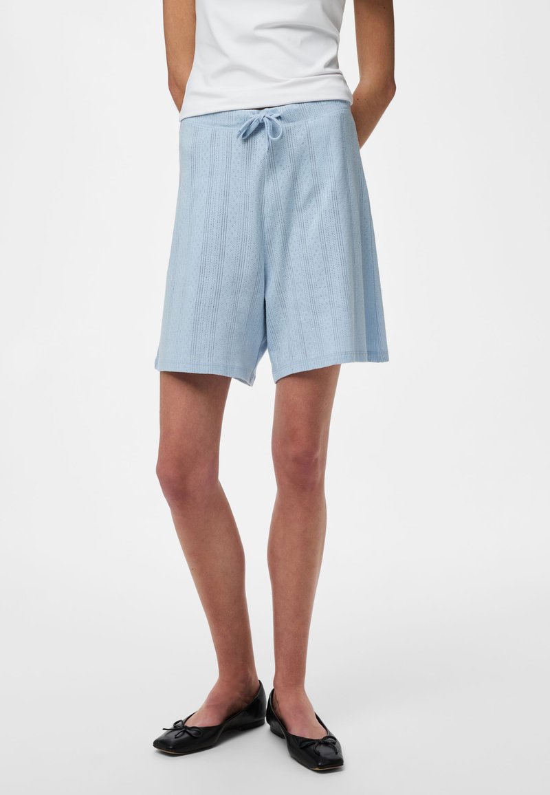 Personne portant un short en maille bleu clair avec une taille à cordon, un haut blanc et des ballerines noires, debout devant un fond uni.