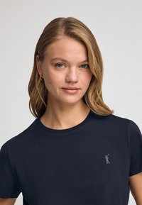 Granatowa koszulka z krótkim rękawem, wykonana z gładkiej bawełny, z małym, srebrnym logo umieszczonym na lewej klatce piersiowej.