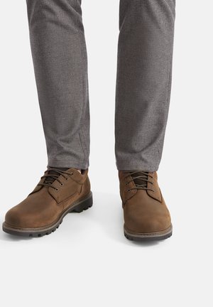 Brązowe skórzane buty z sznurowaniem, zaokrąglonym noskiem i fakturowaną powierzchnią. Połączone z szarymi spodniami w kratę.