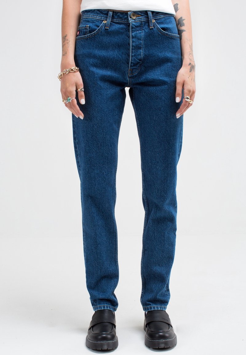 Jeans de mezclilla azul con corte recto, cintura alta, diseño de cinco bolsillos y cierre de botón dorado, combinados con zapatos negros sin cordones.