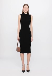 Rochie midi ajustată, neagră, din material răsucit, cu gât înalt și fără mâneci, asortată cu o geantă bej și pantofi negri cu vârf ascuțit.