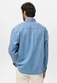 Ljusblå denimskjorta med rygghölje, långa ärmar och rundad fåll. Har diskret sömnad och knäppta manschetter.