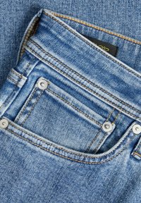 Jeans de mezclilla con un lavado azul claro, que presentan remaches de latón, un pequeño bolsillo delantero y costuras naranjas a lo largo de las costuras.