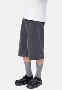 Person wearing long black shorts, gray ribbed socks, and black sneakers with patterned laces on a plain white background.