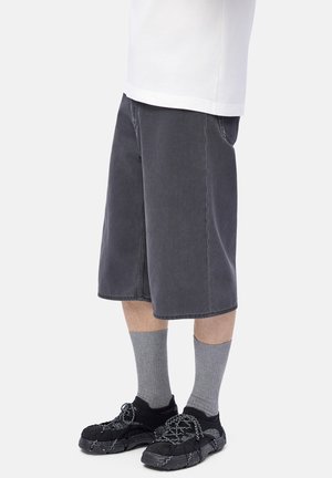 Person som bär långa svarta shorts, grå ribbad strumpor och svarta sneakers med mönstrade snörningar på en enfärgad vit bakgrund.