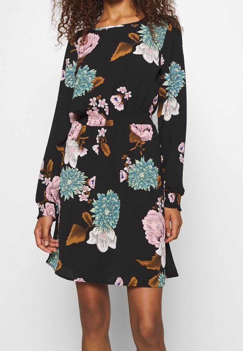 Femme portant une robe noire à manches longues avec des motifs floraux bleus, roses et blancs, une taille élastique et des poignets froncés et volantés.
