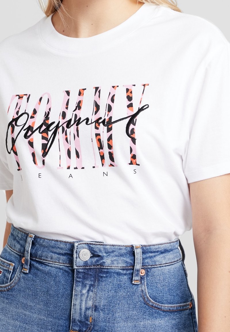 Weißes Baumwoll-T-Shirt mit auffälligem schwarzen und pinken "TOMMY ORIGINAL"-Schriftzug in einem Leopardenmuster. Kombiniert mit hoch geschnittenen blauen Jeans.