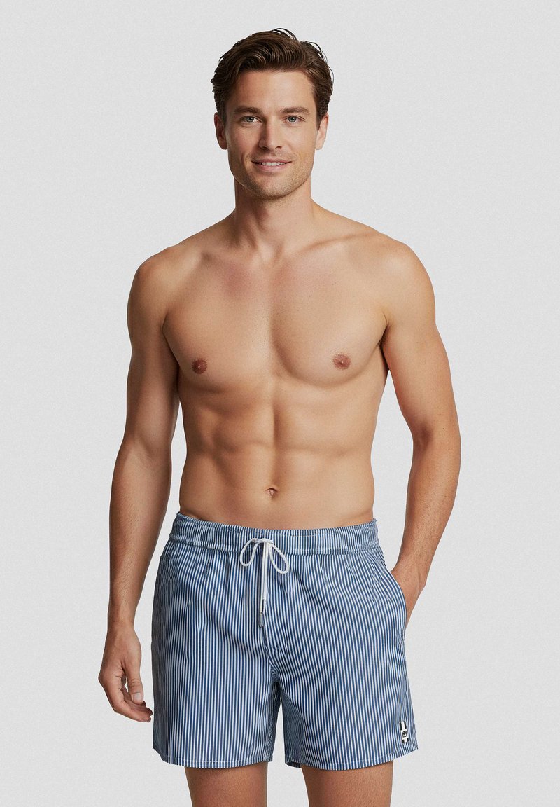 Uomo senza maglietta con capelli castani corti che indossa pantaloncini da nuoto a righe verticali blu e bianche con coulisse bianca, in piedi contro uno sfondo neutro.