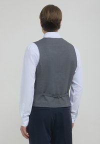 Mann in einem weißen Hemd, grauem Weste mit schwarzem Riemen und dunkler Hose, der mit dem Rücken zu einem schlichten hellen Hintergrund steht.