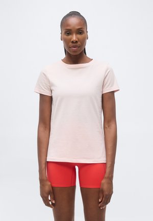 Femeie cu piele închisă la culoare și păr drept, purtând un tricou roz pal cu mâneci scurte și pantaloni scurți fitted roșii aprinși, stând în picioare în fața unui fundal alb simplu.