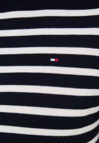 Svetr s námořnickými a bílými pruhy vyrobený z pleteného materiálu, s malým logem v červené, bílé a modré barvě na levé straně.