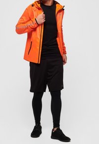 Personne portant une veste orange sur une chemise noire, un short noir, desleggings noirs et des baskets noires sur un fond blanc uni.