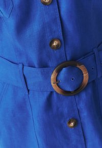 Robe en lin bleu avec des boutons marron et une large ceinture ornée d'une boucle ronde en écaille de tortue.