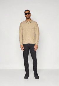 Beežist nööbitav särk rinnataskutega, sobitatud mustade kitsaste pükste ja mustade tossudega. Modell kannab tumedaid päikeseprille, seisab heleda tausta ees.