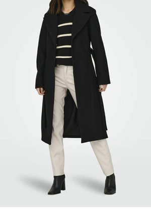 Femme portant un long manteau noir par-dessus un pull rayé noir et blanc, un pantalon beige et des bottines à talons noires, debout les jambes écartées.