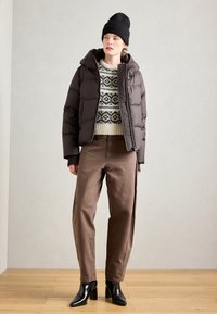 Doudoune marron foncé en tissu texturé avec une capuche, associée à un pull en maille présentant un motif noir et crème, et un pantalon marron.