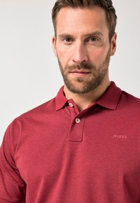 Bordová polo tričko s límečkem, dvěma knoflíky a decentním logem "JP1880" na hrudi. Měkká textura s jemným melírováním.