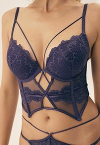 Fioletowy body z koronką i przezroczym paneliem siateczkowym. Posiada krzyżujące się ramiączka i kwiatowe wzory wzdłuż brzegów oraz na środku, co dodaje finezyjnych detali.
