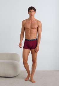 Boxeri burgundy cu un brâu negru cu model, având o croială strânsă și o textură fină, purtați de un model într-un mediu neutru.