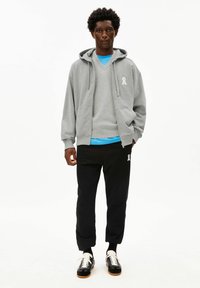 Grijze hoodie met rits over een blauw T-shirt, gecombineerd met zwarte sweatpants met een wit lintlogo en zwart-witte sneakers.